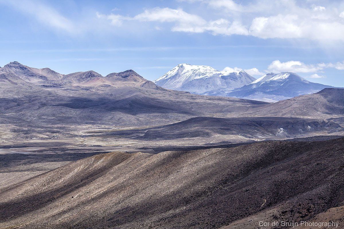 Barren mountain range under a cloudy blue sky. Snow-capped peaks in the distance.