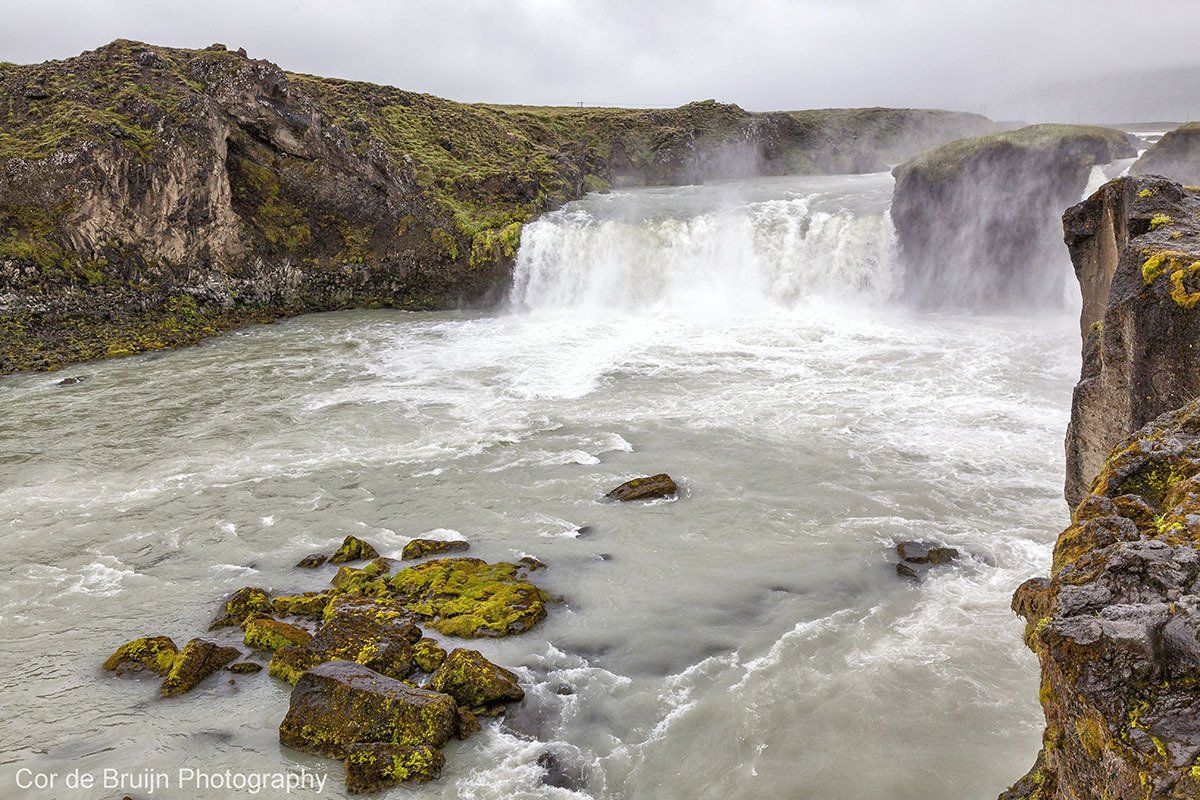 Waterfall cascading into a river with mossy rocks and overcast sky.
