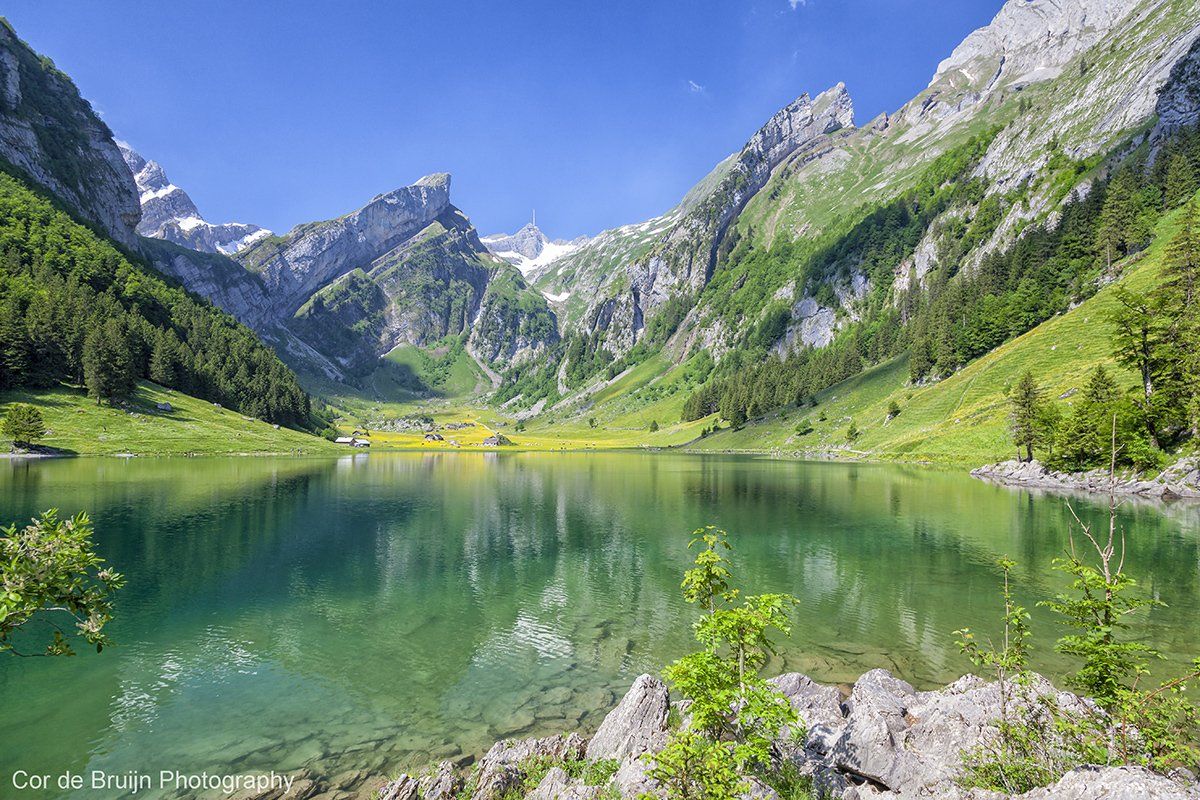 Crystal-clear alpine lake reflects rugged mountain peaks and lush green meadows under a bright blue sky.