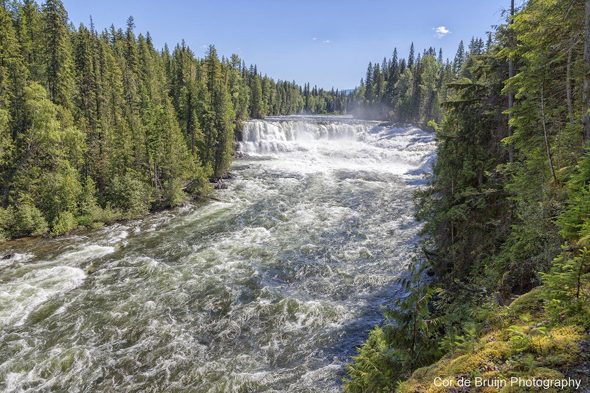 Water cascades over tiered falls surrounded by a dense green forest, under a clear blue sky.