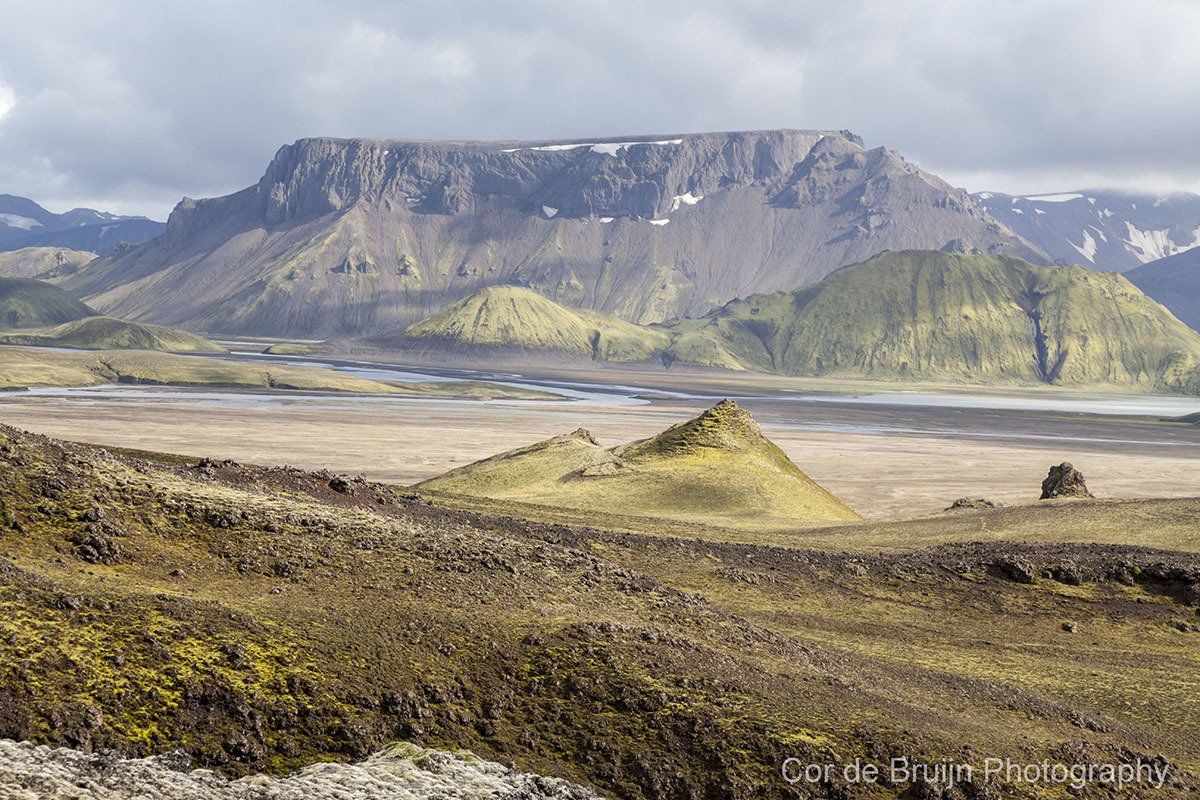 Landscape of a rugged, flat-topped mountain with snow, green hills, and a vast, muted-colored plain.