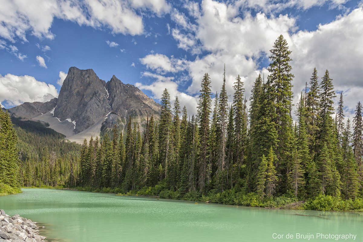 Emerald-green lake in front of tall evergreen trees; jagged mountain peaks in the background, blue sky with clouds.