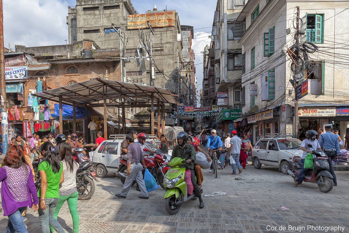 Busy street in Kathmandu, Nepal, with traffic and pedestrians, lined with shops and buildings.