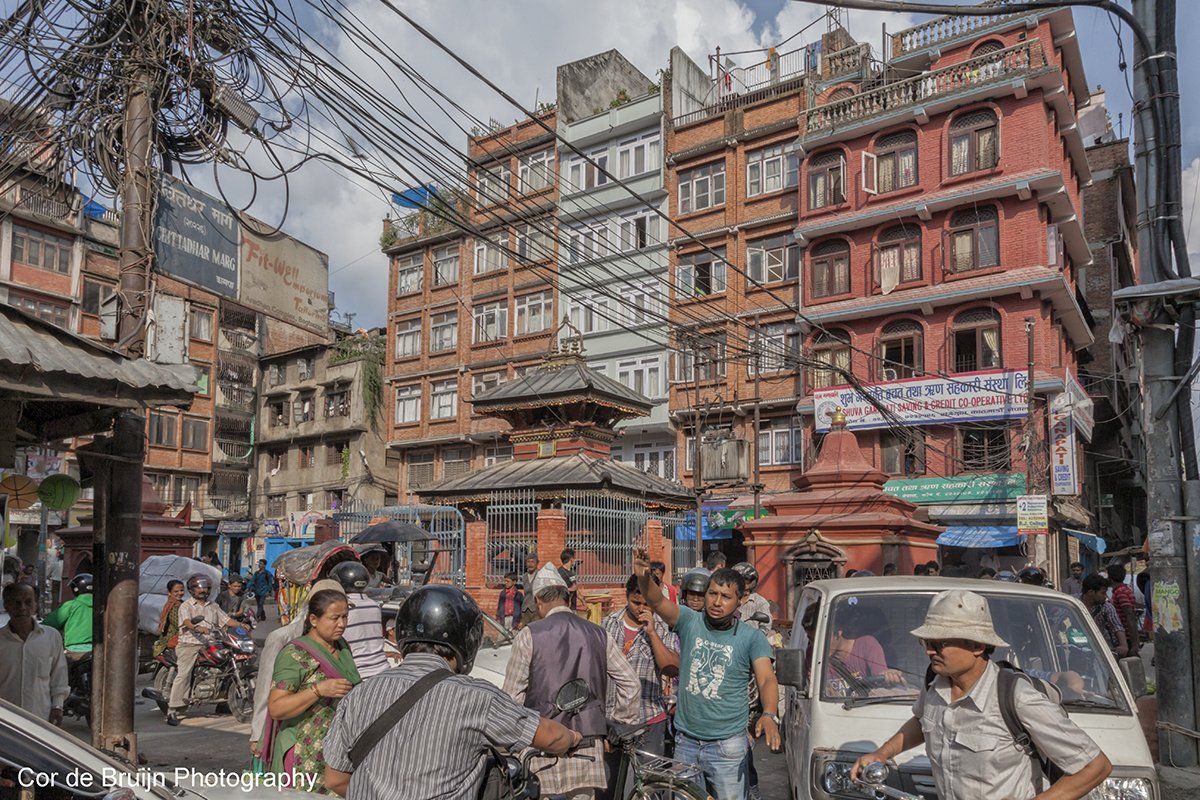 Busy street scene in Kathmandu, Nepal, with buildings, people, vehicles, and tangled overhead wires.