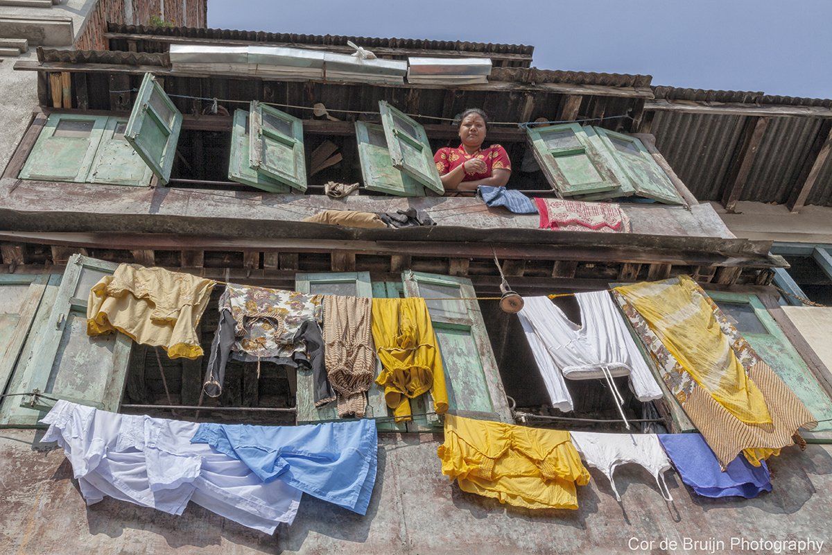 Woman leans from a window above laundry drying outside in a weathered, multi-windowed building.