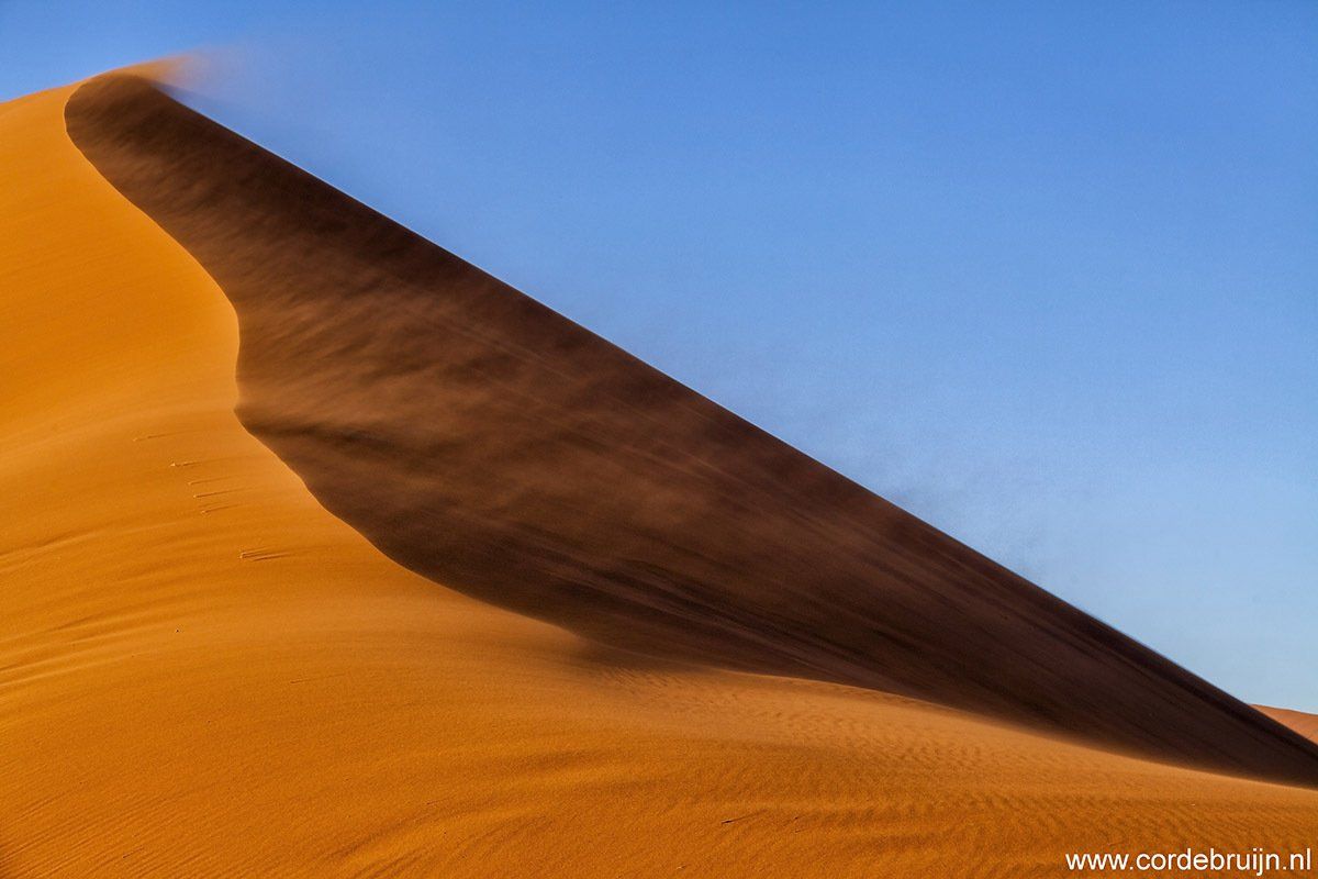 Sand dune with wind blowing sand against a bright blue sky.