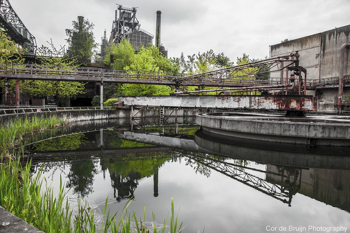 Abandoned industrial complex with water basins reflecting structures, overgrown with vegetation, overcast sky.