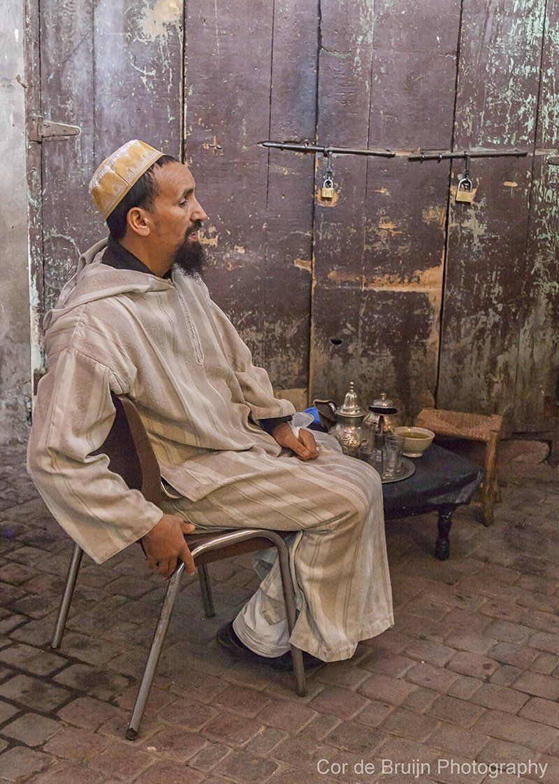 Man in traditional attire sits outside, leaning against a weathered wooden door.