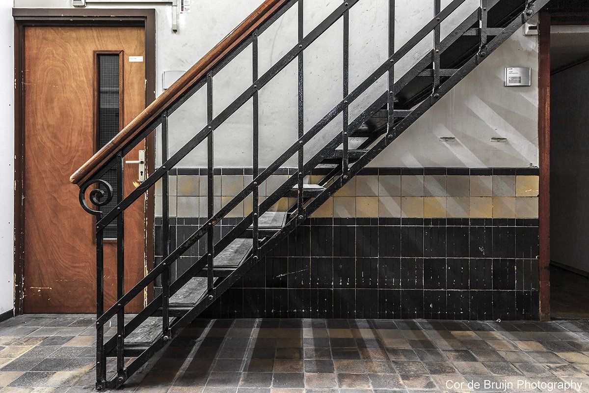 Staircase with black metal railing and wooden handrail against tiled wall and wooden door.