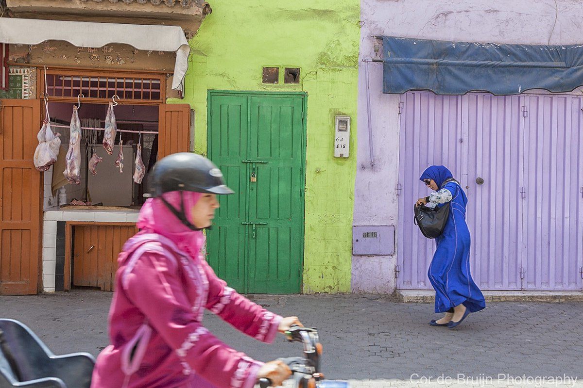Woman in pink riding a scooter, another woman in blue carrying a bag on a colorful street with shops.