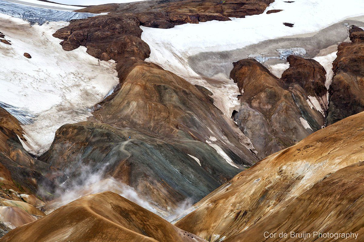Colorful mountain landscape with snow patches, steam, and varying shades of brown and green.