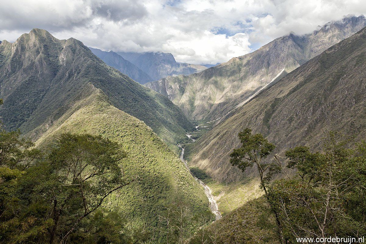 Mountain valley with a river winding through, lush green slopes, and cloudy sky.