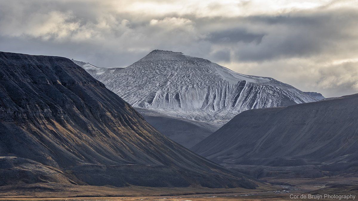 Snow-covered mountain peak rises above dark, ridged valleys under a cloudy sky.