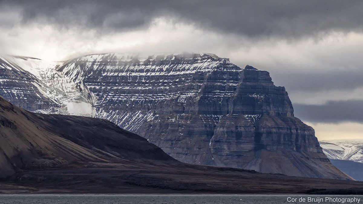 Snow-capped mountain under cloudy sky, with dark, layered rock face and low-lying dark land in the foreground.