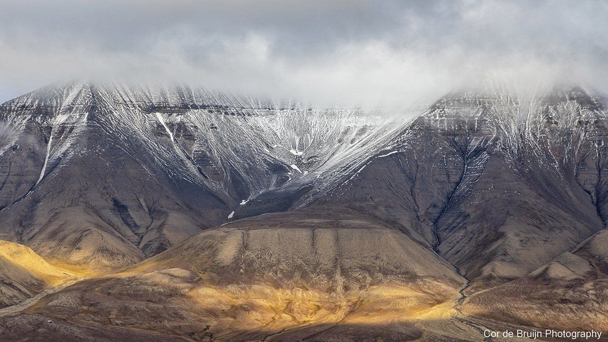 Snow-dusted mountain range under cloudy sky. Shadows and sunlight create contrast on the slopes.