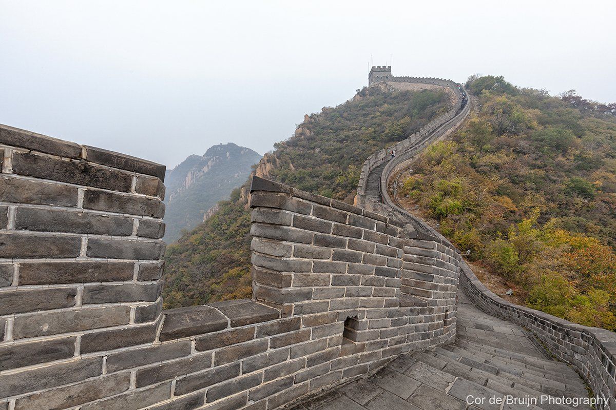 Stone wall of the Great Wall of China winds along a ridge with steps, under a hazy sky.