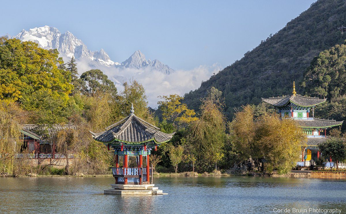 Pavilion on lake with snow-capped mountain backdrop, trees, and traditional Chinese architecture.