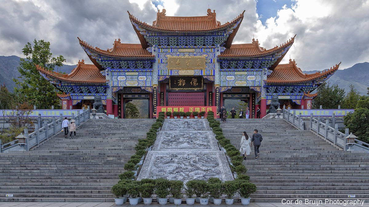 Ornate Chinese temple entrance with people walking on stairs. Blue, red, and gold accents, mountains in the background.