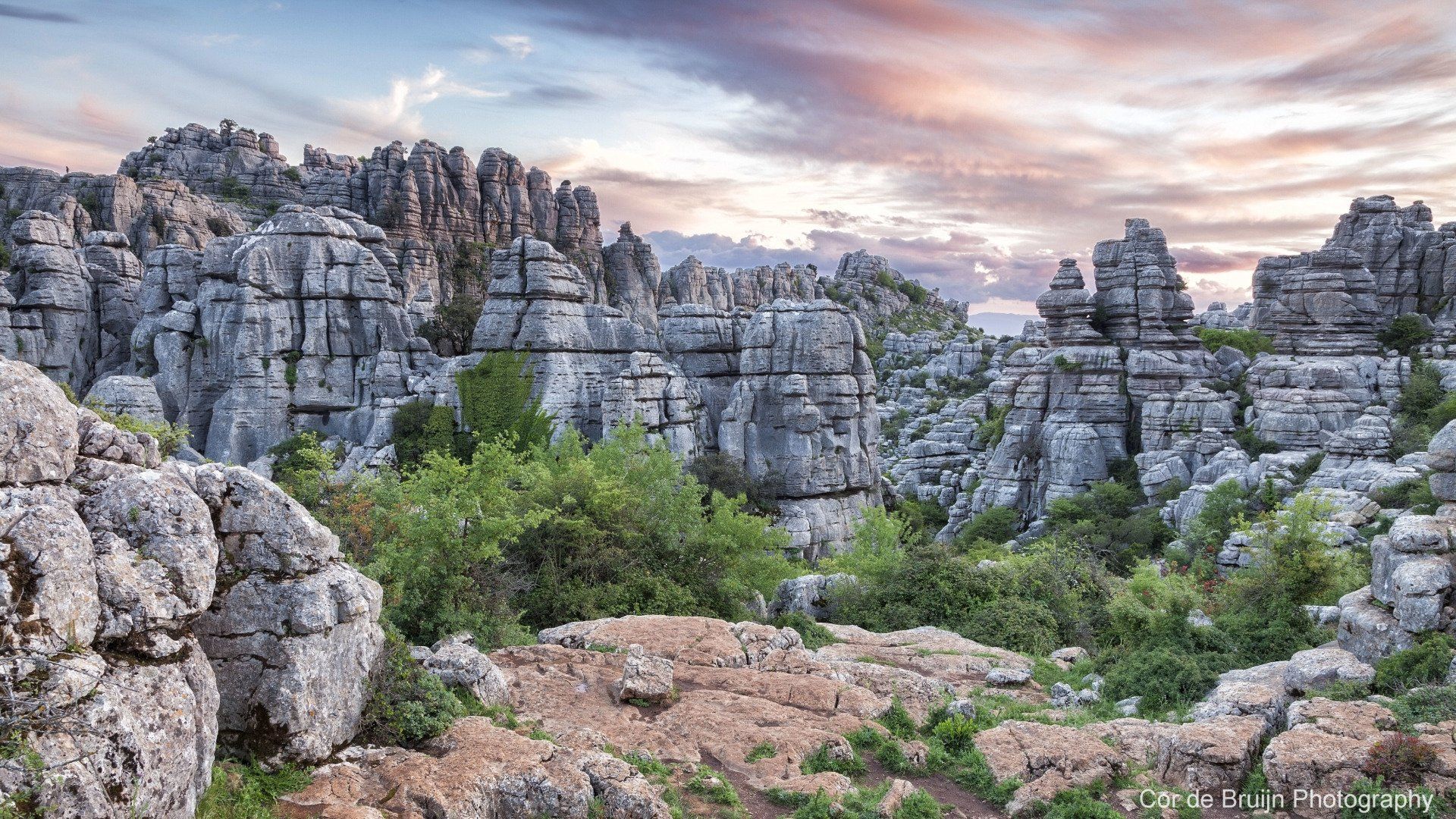 Rocky, gray limestone formations with green vegetation under a colorful sunset sky.