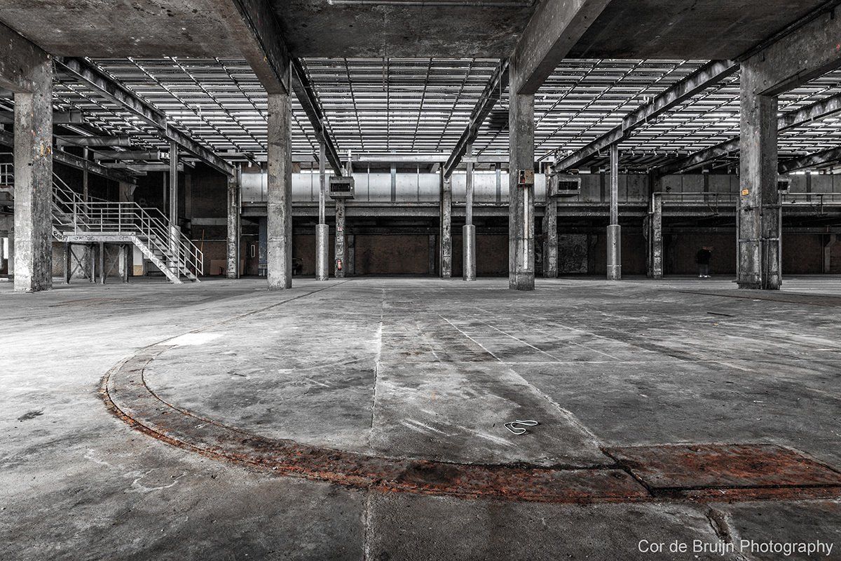 Interior view of an abandoned industrial building. Concrete floors, support pillars, and metal framework with dim lighting.