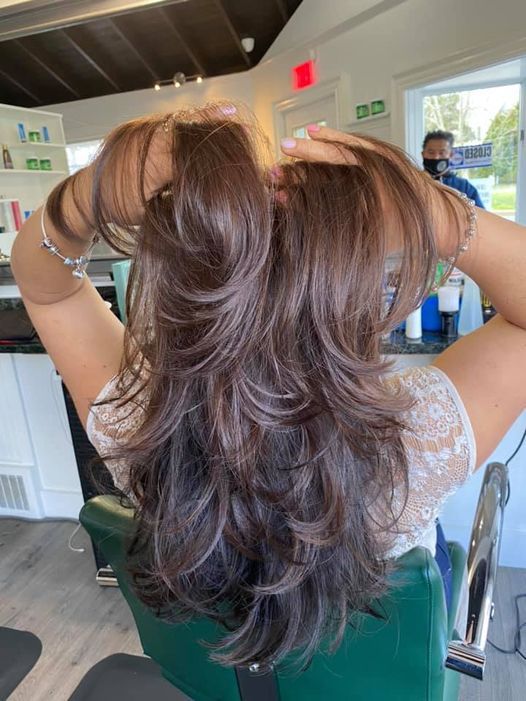 Person with layered brown hair raising both arms in a salon chair, viewed from behind.