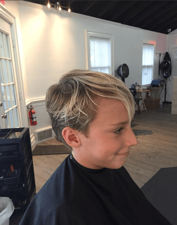 Side-profile of a person with short blond hair seated in a salon, wearing a black cape and faintly smiling.