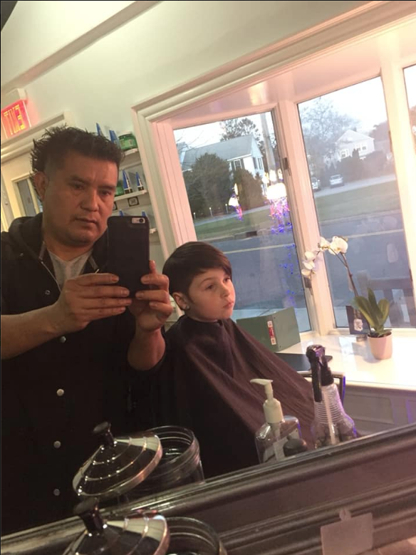 Man and child in a salon mirror selfie, with a window and city view behind them.