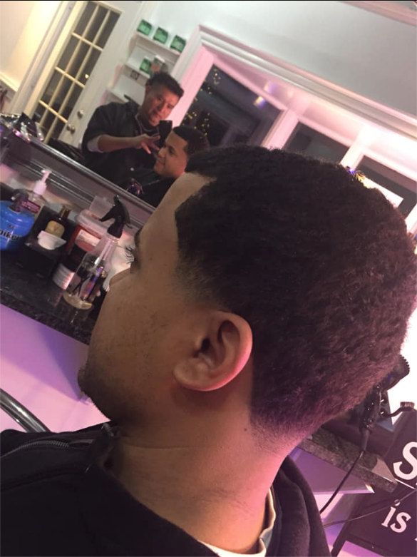 Person with a short haircut sits in a salon chair, looking toward a mirror and holding a phone.