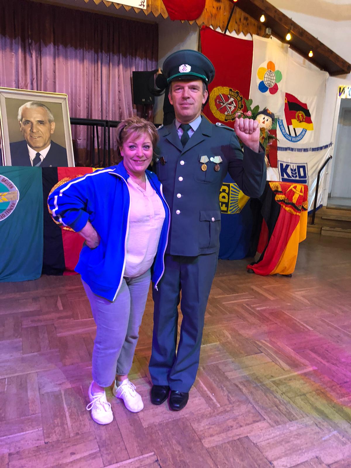 Zwei Personen posieren im Innenbereich. Mann in Uniform, Frau im Trainingsanzug, Flaggen und Porträt im Hintergrund.