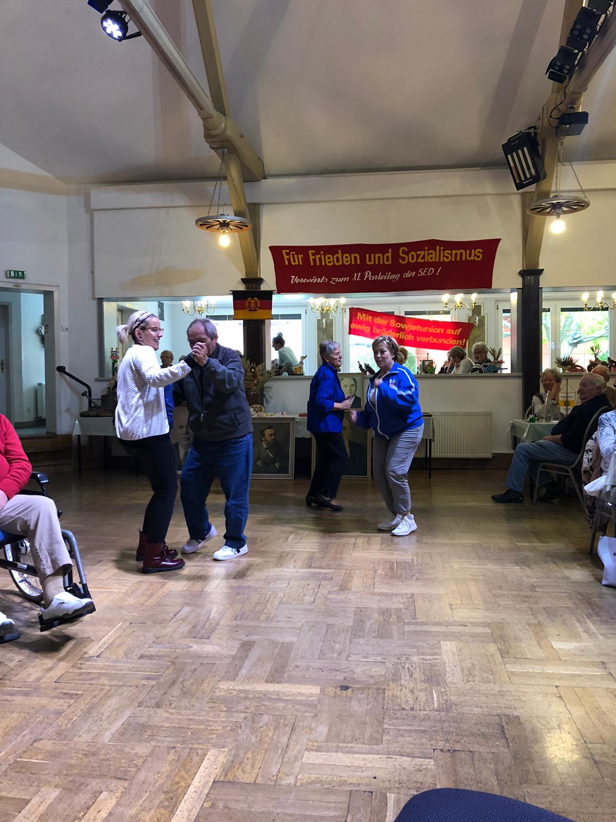 Menschen tanzen in einem Saal. Mann in Jeansjacke tanzt mit einer Frau. Andere schauen zu, rotes Banner.
