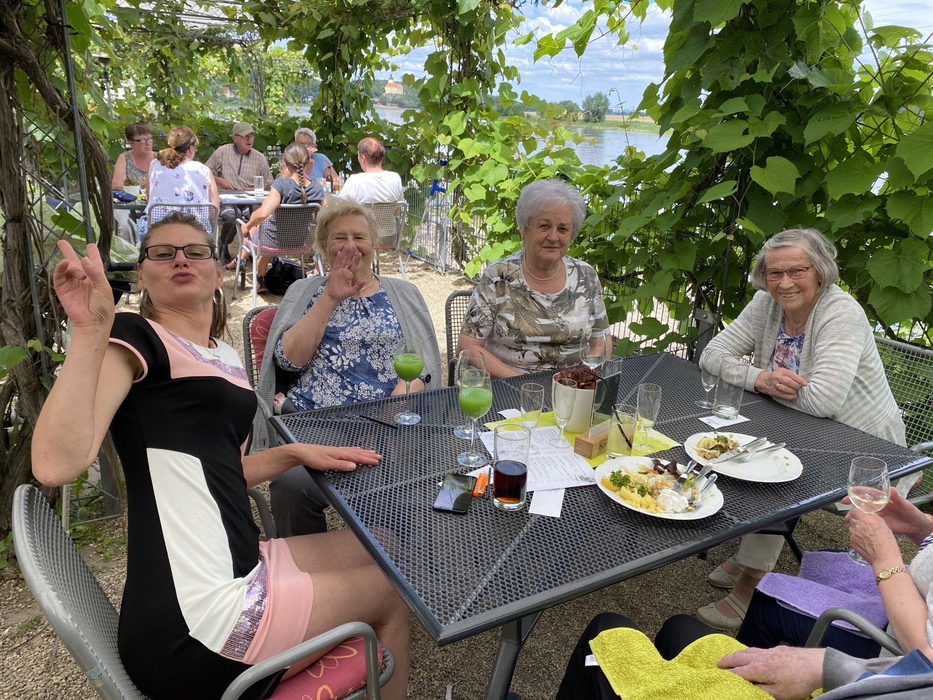 Eine Gruppe von Menschen sitzt an einem Tisch im Freien unter einer weinbewachsenen Pergola und trinkt und isst.