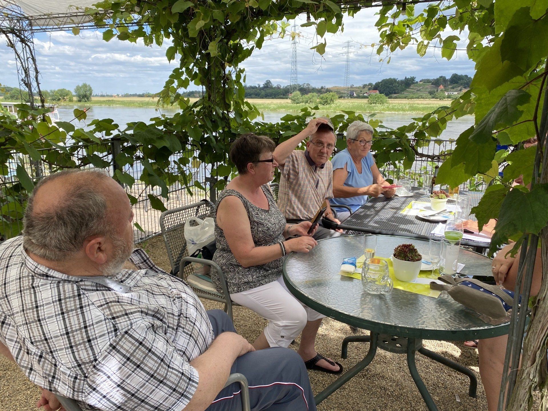 Eine Gruppe von Menschen sitzt an einem Tisch unter einer weinbewachsenen Laube mit Blick aufs Wasser.