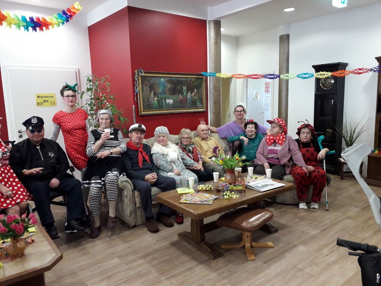Eine Gruppe von Menschen in Kostümen sitzt in einem Wohnzimmer, einige lächeln und feiern mit Dekorationen.