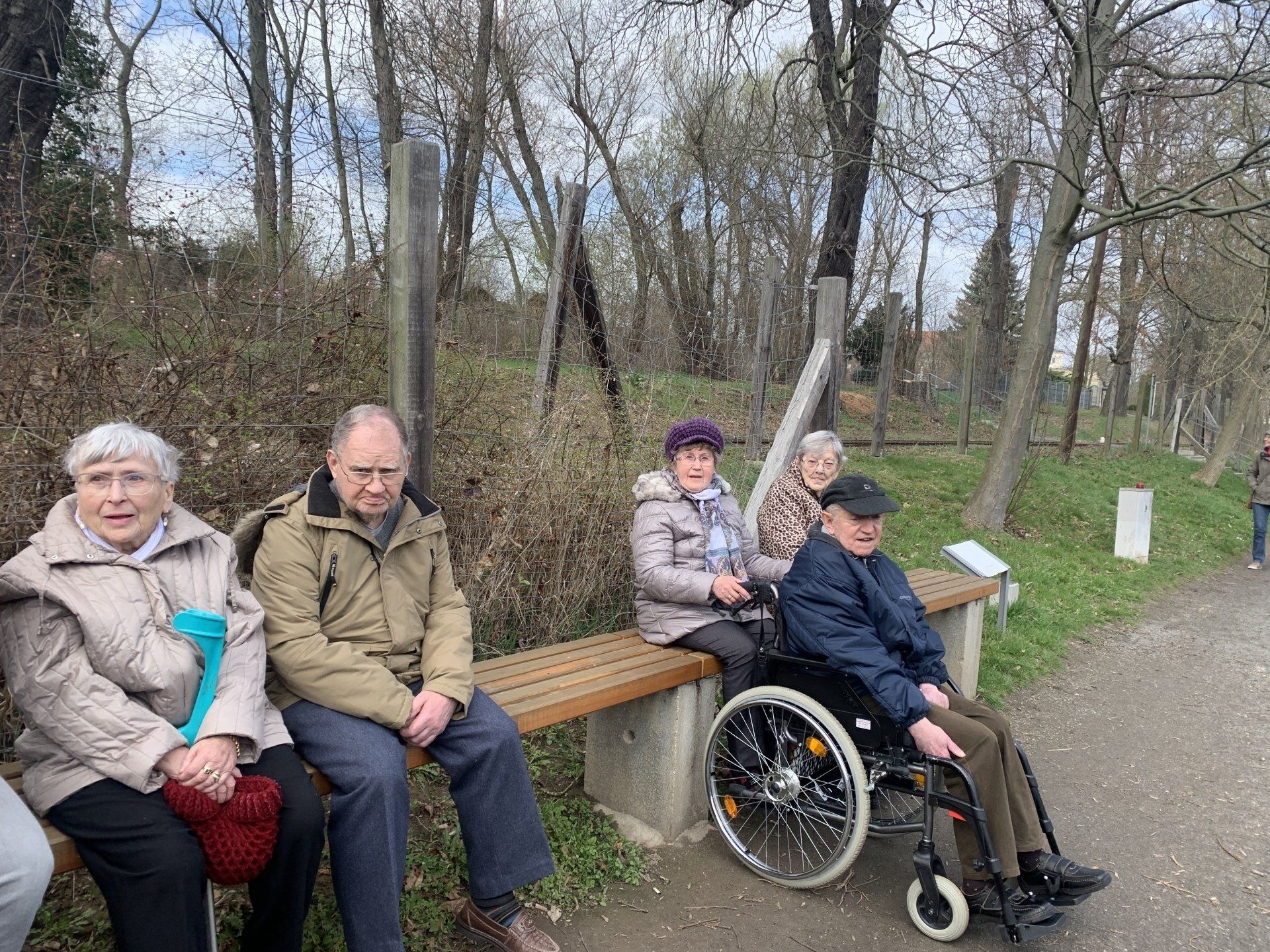 Fünf Personen sitzen auf einer Parkbank. Eine Person sitzt im Rollstuhl. Im Hintergrund sind Bäume und ein Weg zu sehen.