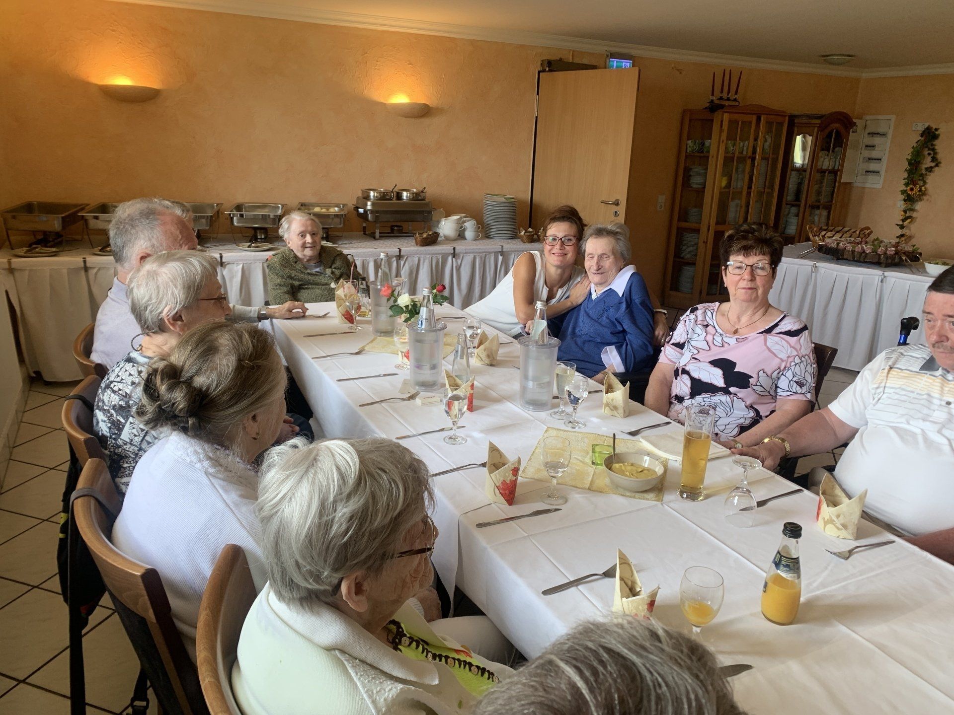 Menschen sitzen an einem langen Tisch, vermutlich in einem Restaurant. Im Hintergrund Buffets, Gläser und Essen.