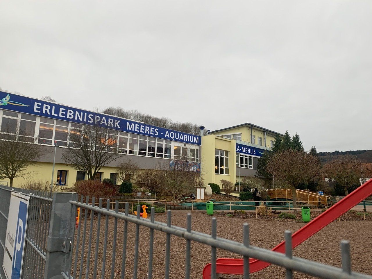 Außenansicht des ErlebnisPark Meeres-Aquarium, ein Gebäude mit blauem Banner, Spielplatz im Vordergrund, bedeckter Himmel.