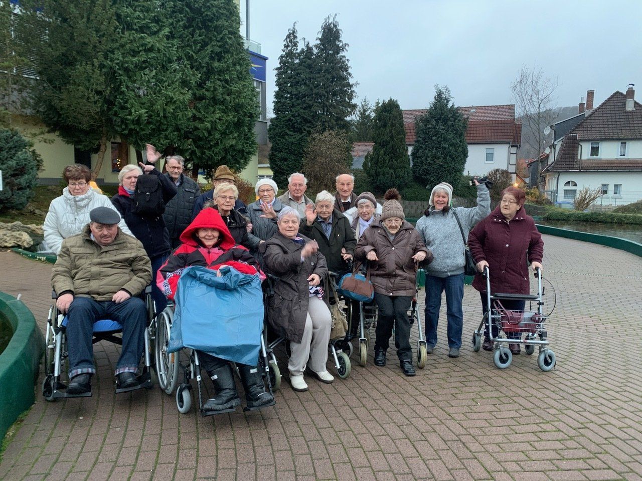 Eine Gruppe von Menschen in Rollstühlen und mit Gehhilfen posiert draußen in der Nähe von Bäumen und Gebäuden.