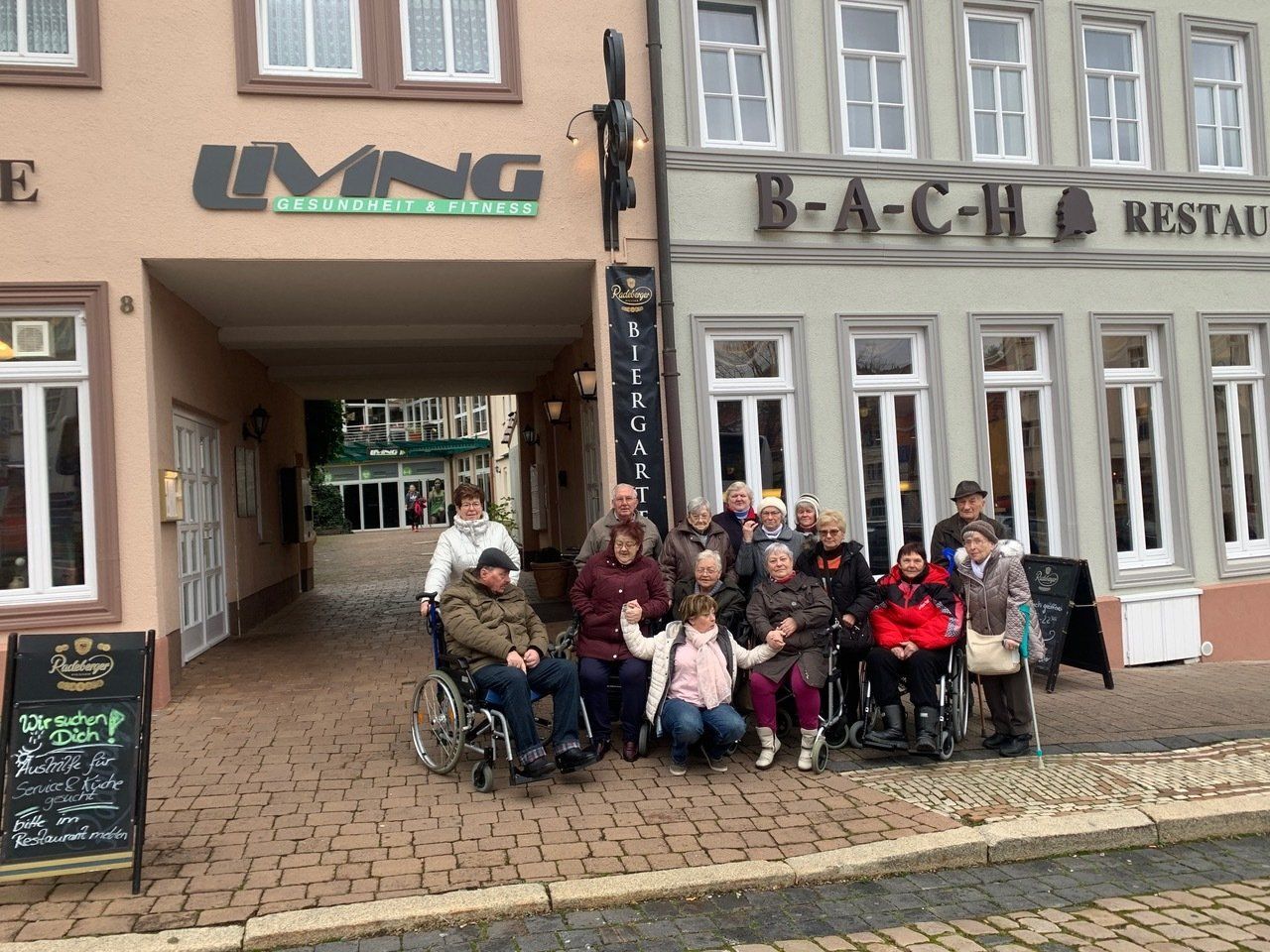Eine Gruppe von Menschen, einige davon im Rollstuhl, posiert vor den Schaufenstern zweier Restaurants: „Living“ und „Bach“.