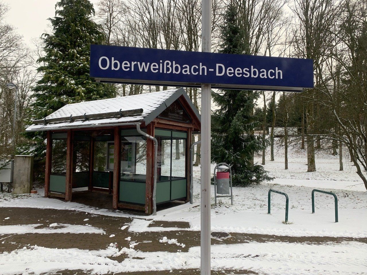 Hinweisschild zum Bahnhof Oberweißbach-Deesbach im Winter. Schnee bedeckt den Boden und einen kleinen Unterstand.