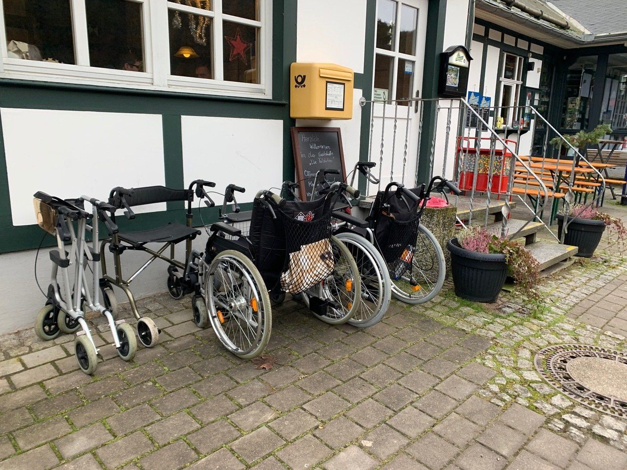 Vor einem Gebäude mit weißer Fassade und grünen Verzierungen stehen mehrere Rollstühle und ein Rollator.