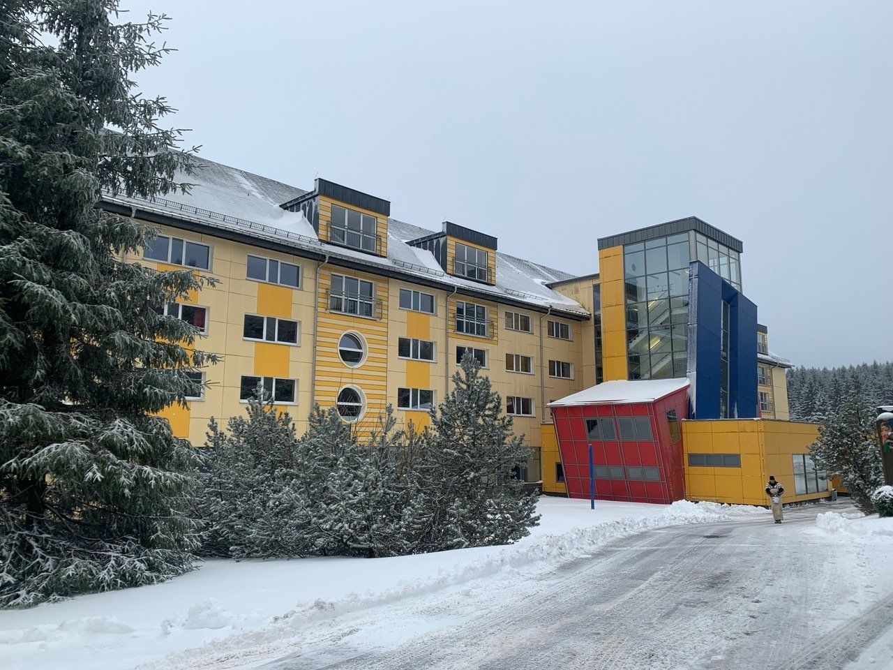 Gelbes Gebäude mit roten und blauen Akzenten, schneebedeckter Boden, immergrüne Bäume.