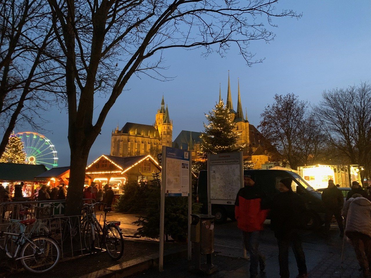 Weihnachtsmarkt in der Abenddämmerung mit Dom im Hintergrund, Riesenrad und beleuchteten Ständen. Menschen sind anwesend.
