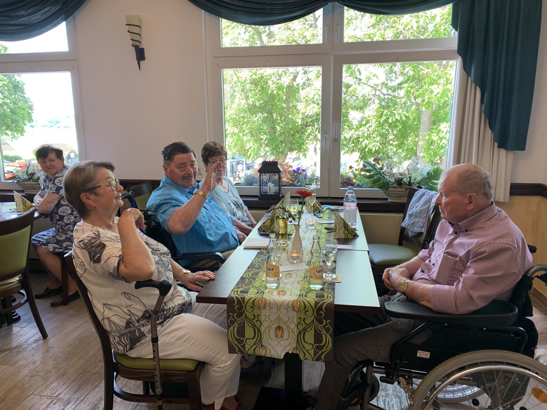 Menschen sitzen an einem Tisch in der Nähe eines Fensters, unterhalten sich und gestikulieren. 