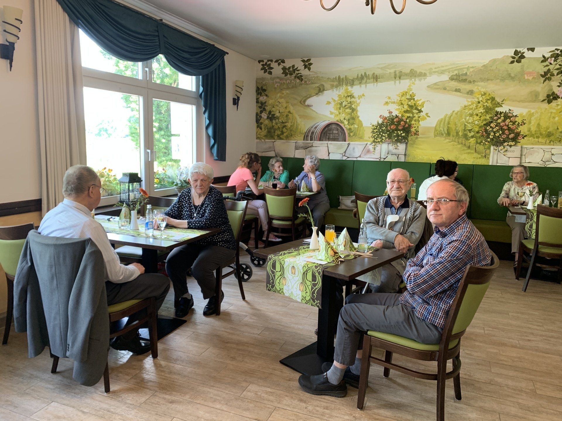 Menschen sitzen an Tischen in einem Café. Einige blicken in die Kamera. An der Wand hängt ein Landschaftsgemälde.