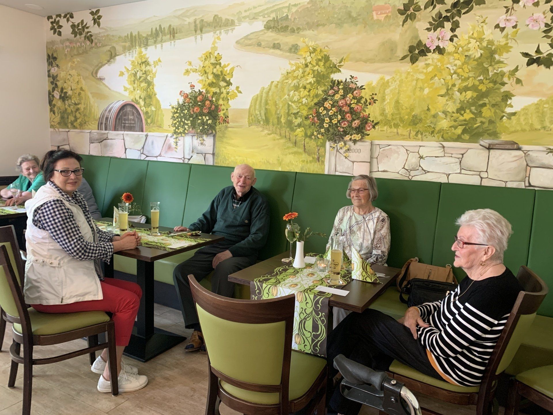 Vier Personen sitzen an einem Tisch in einem Café, an dessen Wand ein Wandgemälde eines Weinbergs hängt.