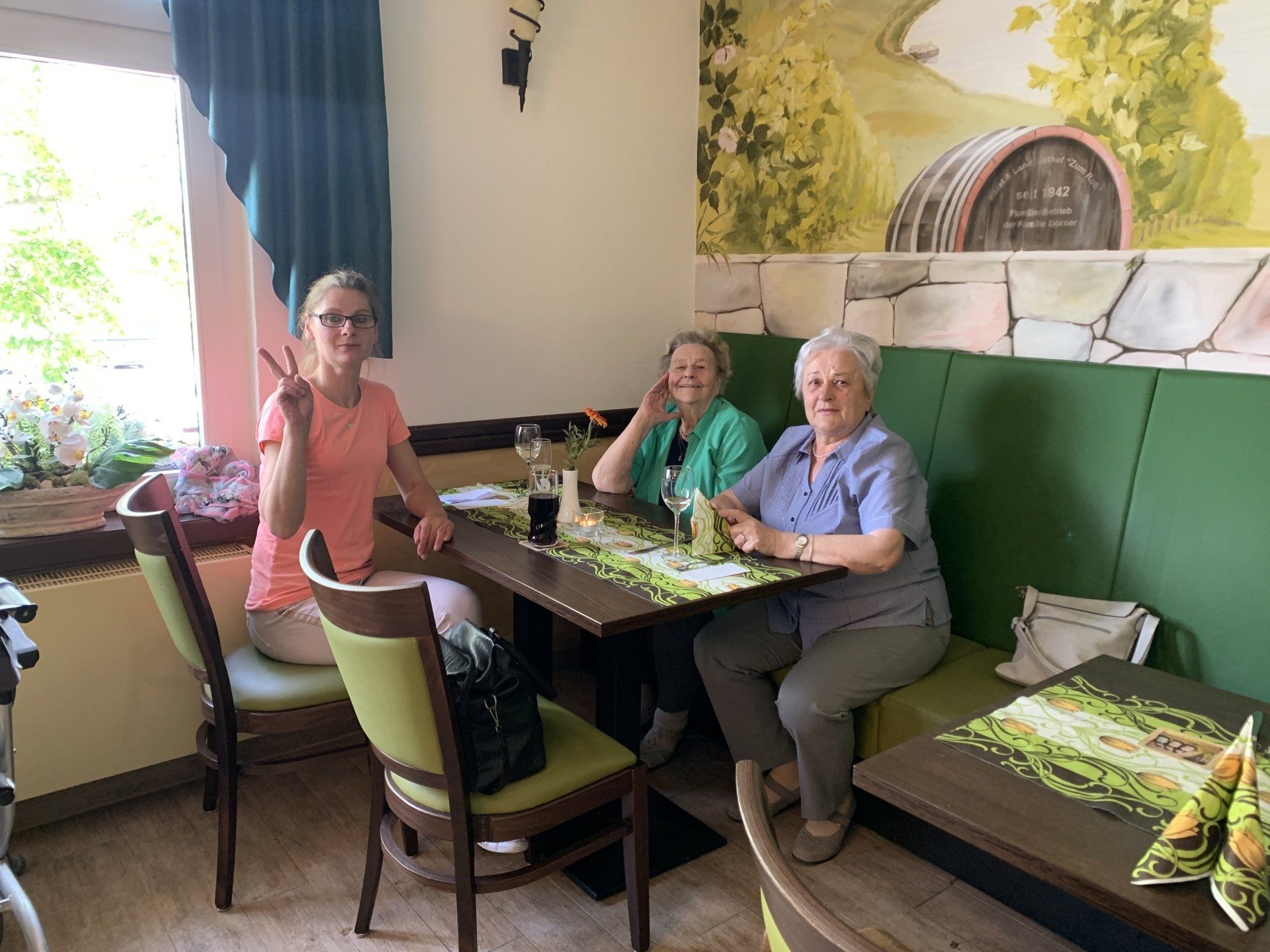 Drei Personen sitzen an einem Tisch in einem Restaurant. Einer macht ein Peace-Zeichen. Blumendekor, eine grüne Sitznische.