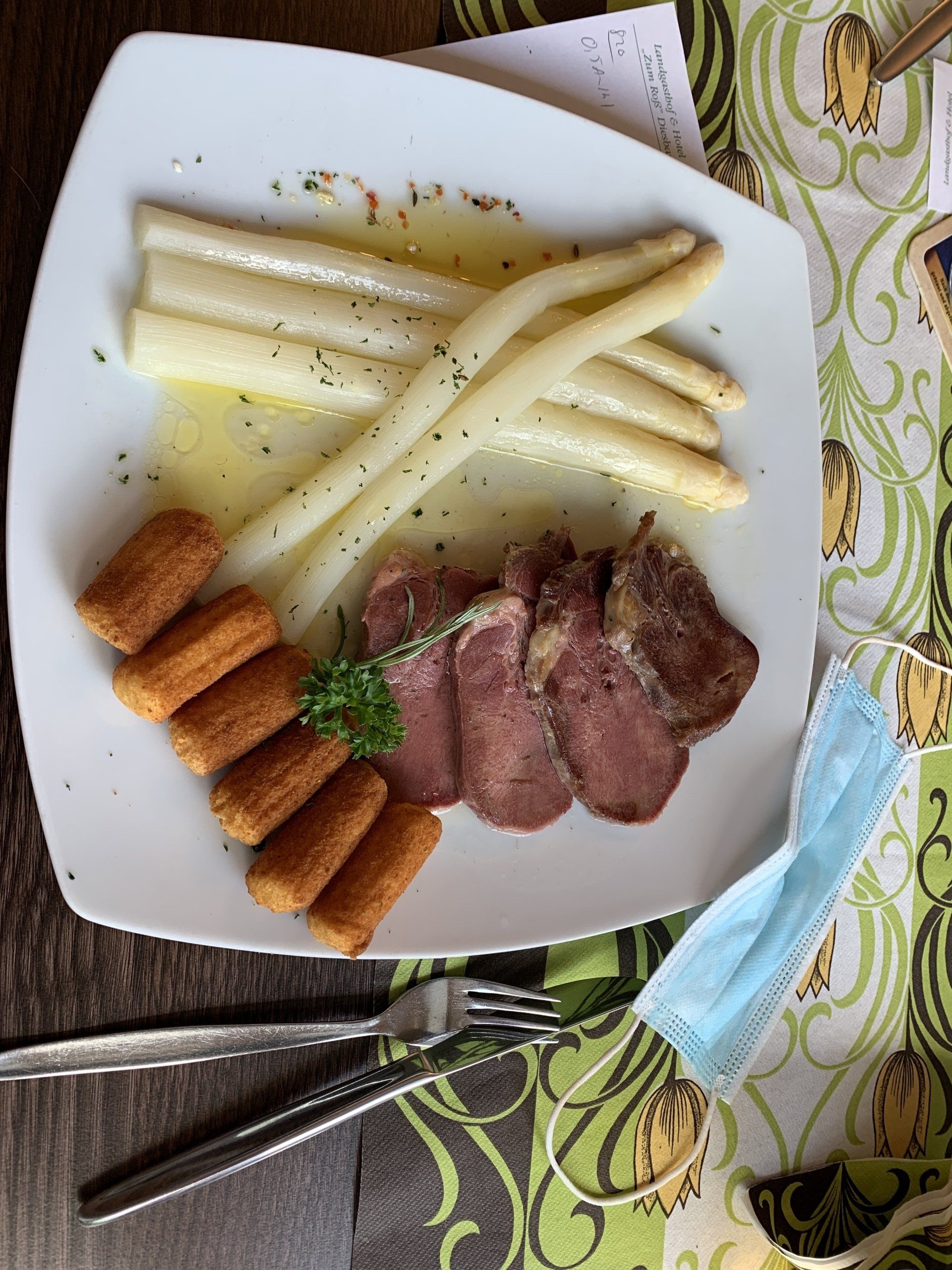 Teller mit weißem Spargel, Fleischscheiben, Kroketten und einer Gesichtsmaske.