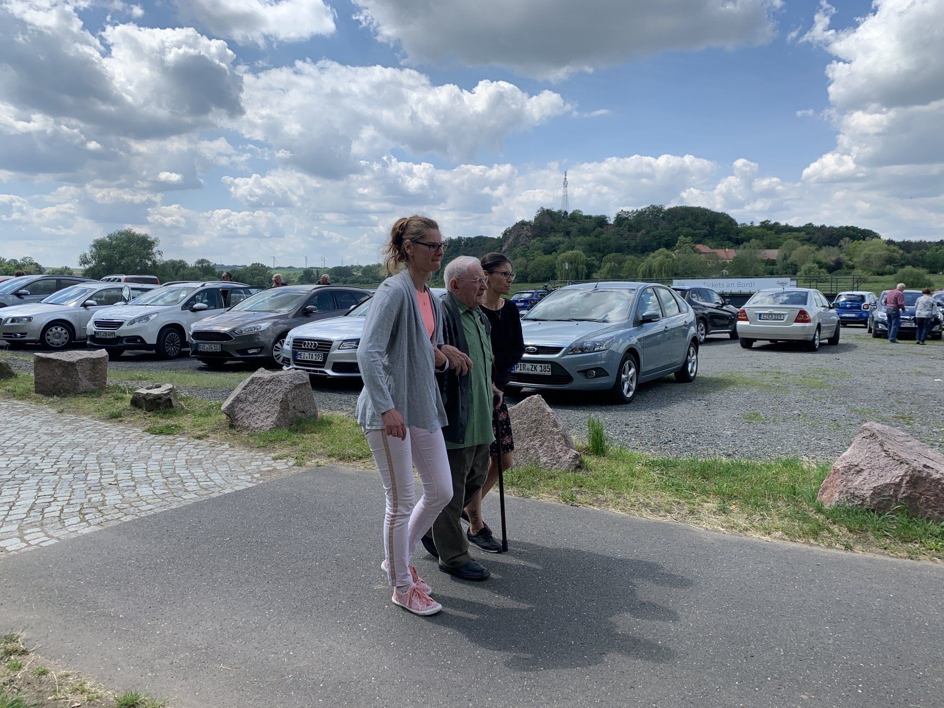 Drei Personen gehen auf einem Parkplatz. Zwei helfen einer Person mit einem Gehstock.