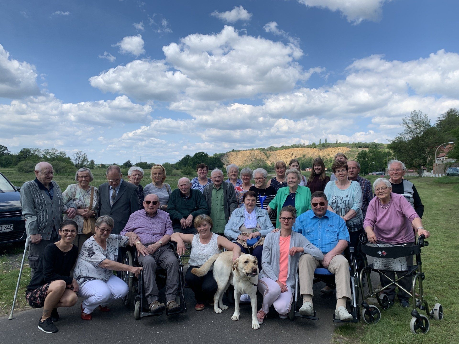Gruppe von Menschen, einige im Rollstuhl, posiert im Freien mit einem Hund. Bewölkter Himmel, Bäume und eine Straße.