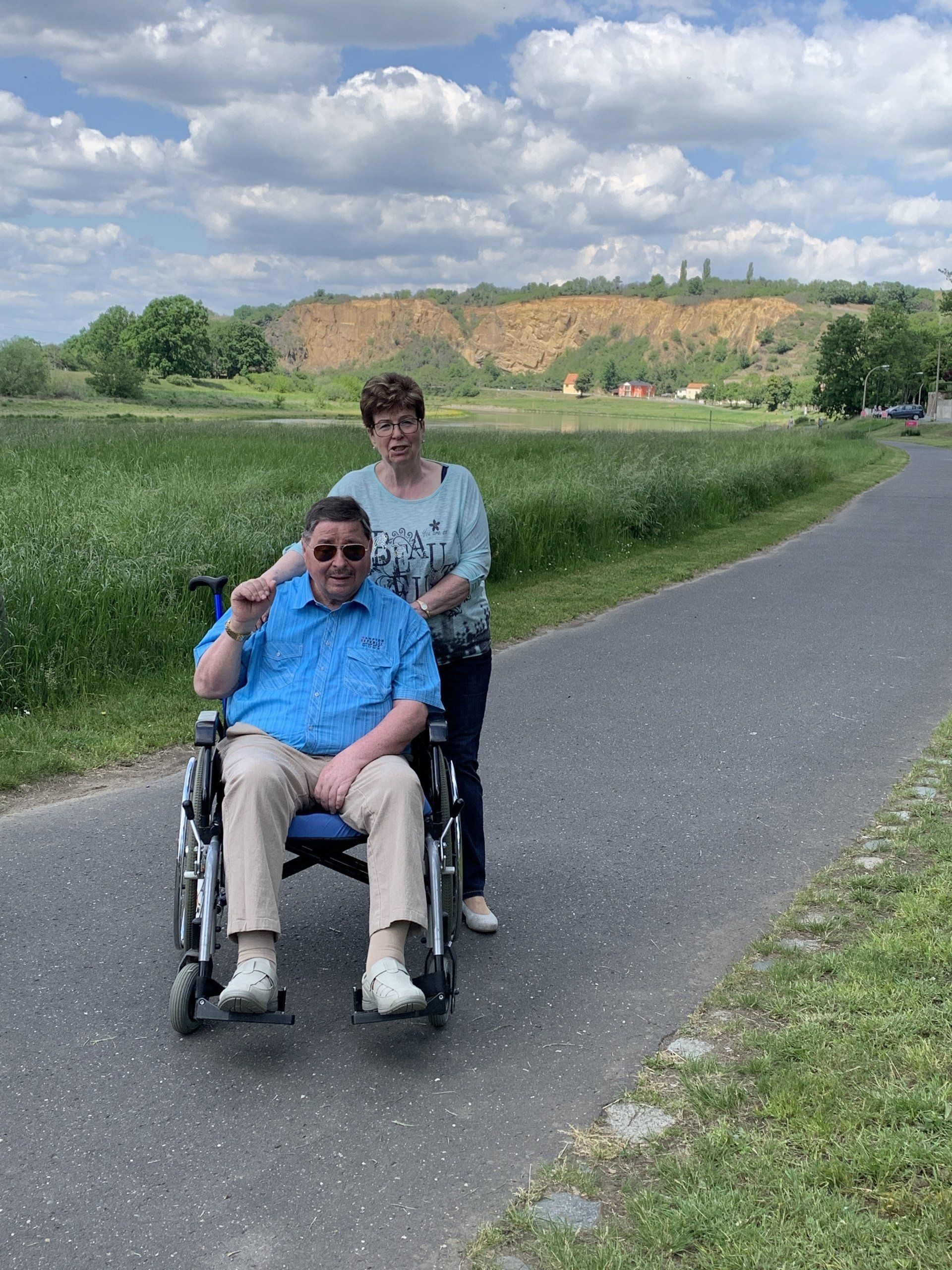Person im Rollstuhl mit Begleiter auf gepflastertem Weg auf einem Feld; sonnig, mit sanften Hügeln im Hintergrund.